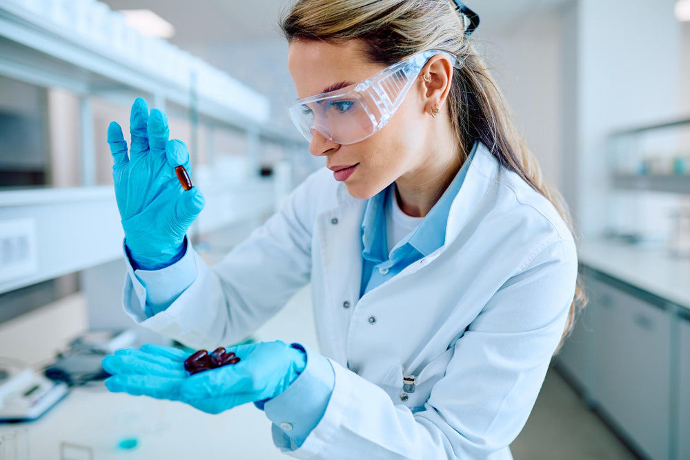 A woman in a white lab coat and blue gloves holds dark capsules in her hand while wearing clear safety goggles in a bright laboratory setting.