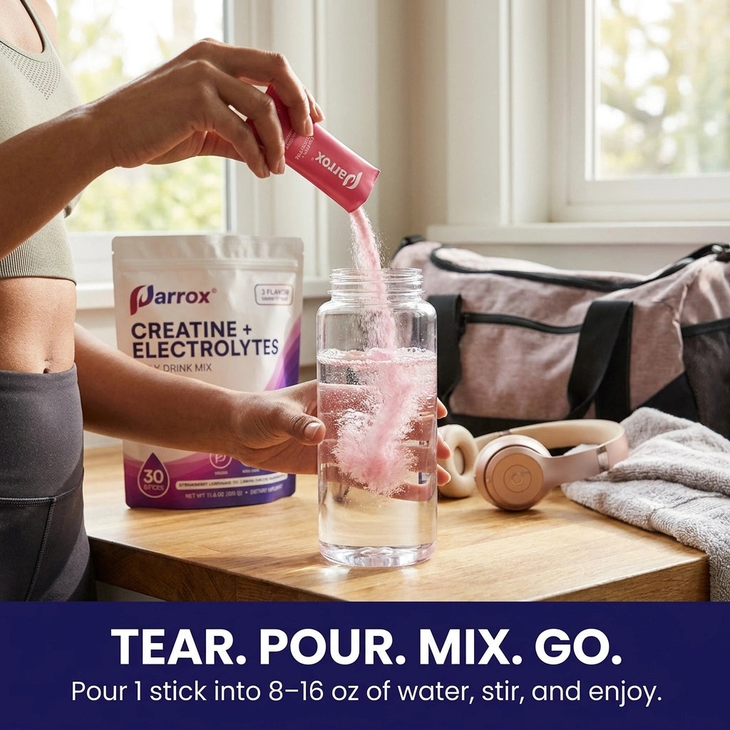 A person pours a pink powder stick into a clear water bottle on a wooden table, with a pouch of Arrox Creatine + Electrolytes and a gym bag in the background near a window.
